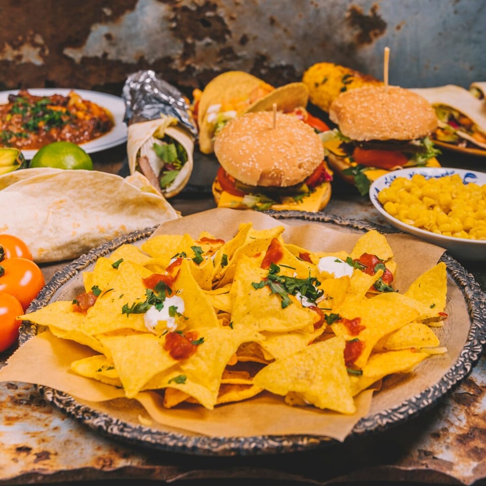 A spread of Mexican food including nachos, burgers, burritos, tacos, and a side of corn.