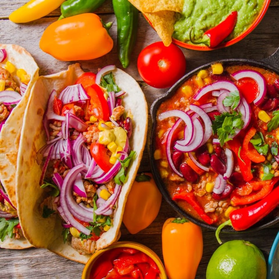 Two tacos with red onion and corn, a skillet of chili, guacamole, and fresh peppers.